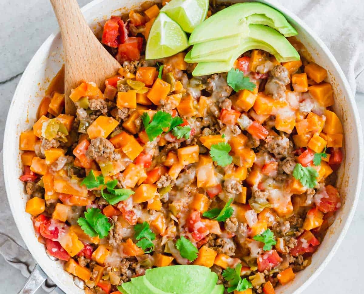 A skillet with cooked ground meat, diced sweet potatoes, tomatoes, cheese, cilantro, and sliced avocado. A wooden spoon is in the pan.
