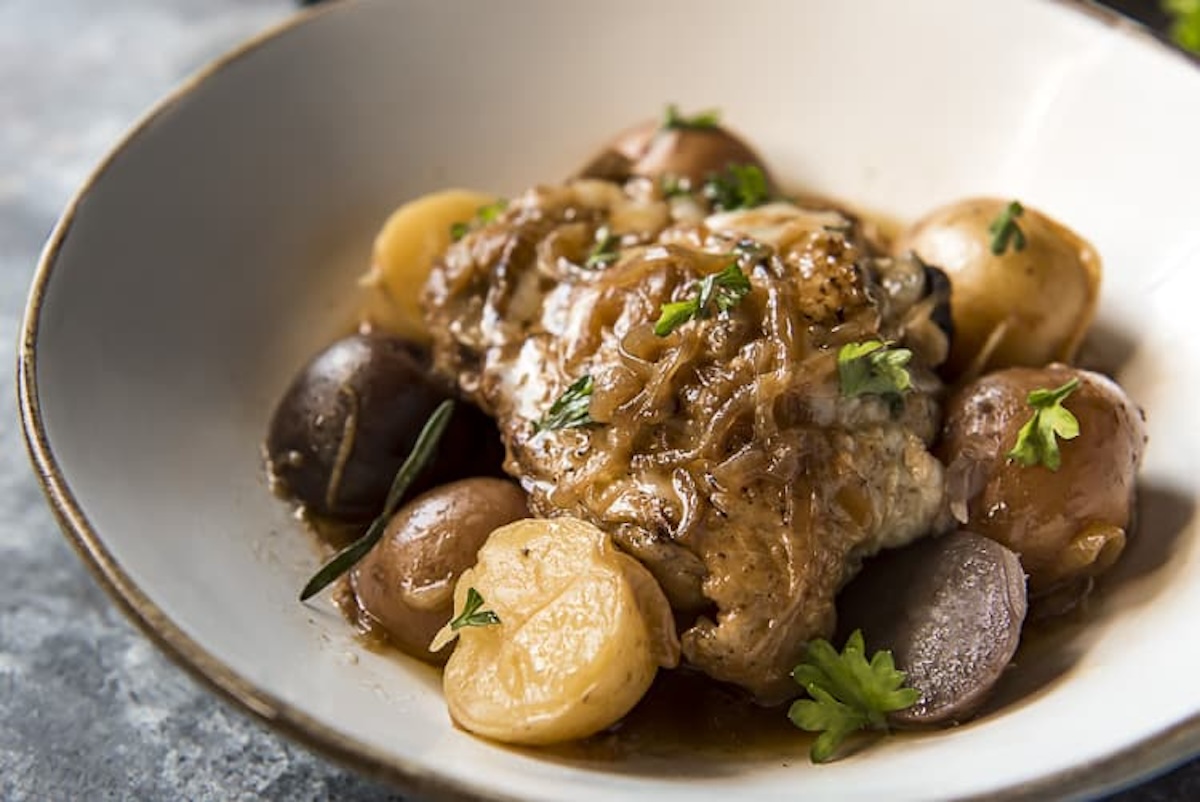 A dish of braised chicken, enhanced with 12 onion pieces, served with various small potatoes and garnished with herbs in a white bowl.