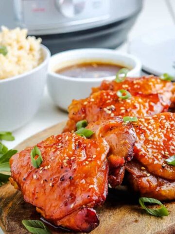 A wooden board with glazed chicken pieces garnished with green onions. A bowl of rice and a small dish of sauce are in the background, along with utensils and a blue-striped napkin.