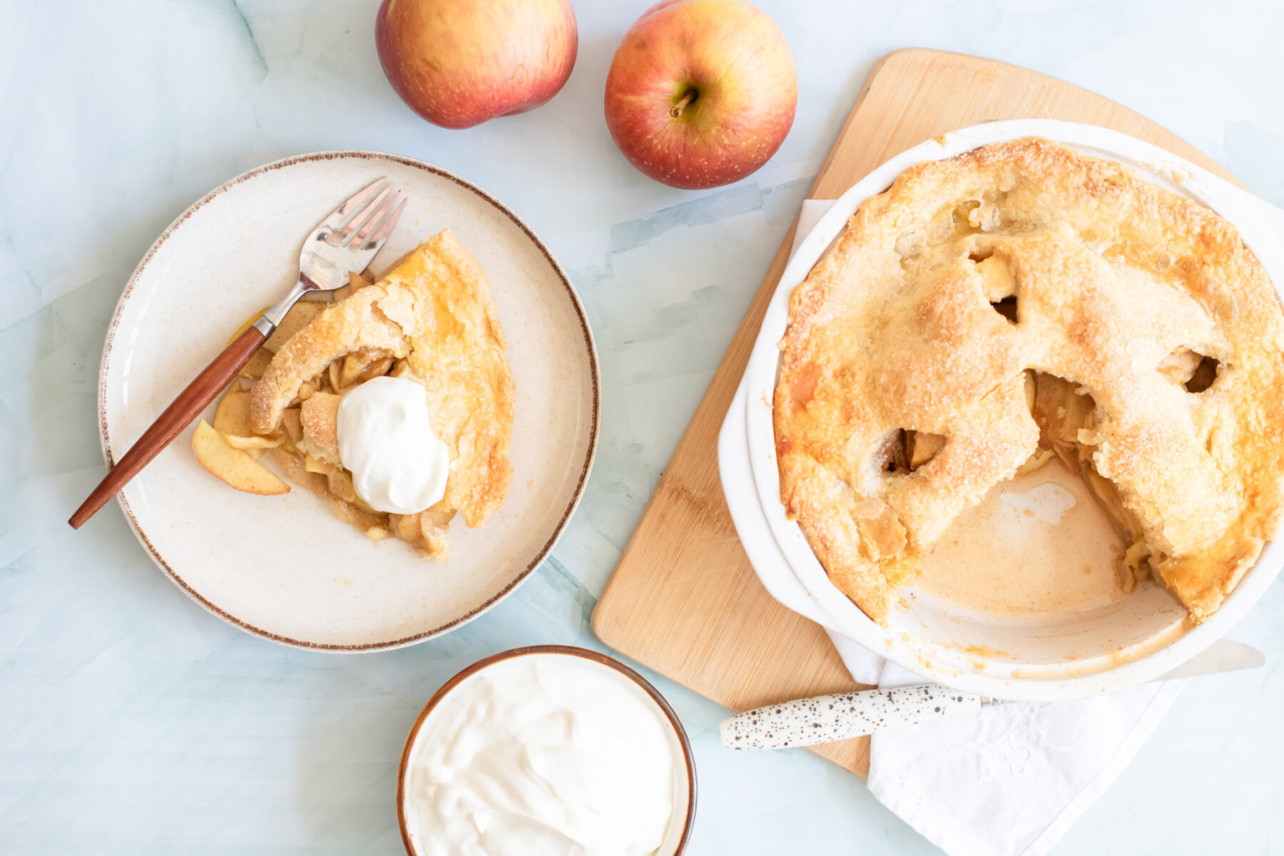 A sliced apple pie with a serving on a plate, topped with cream. Two apples and a bowl of cream are nearby on a light background.