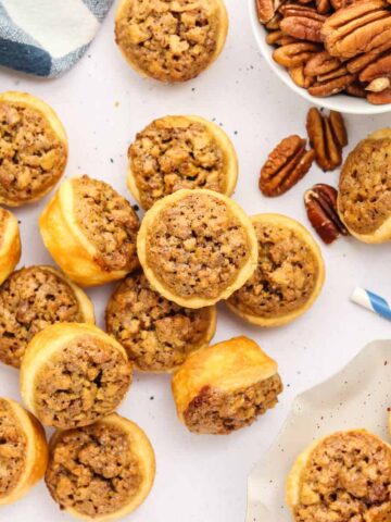 Mini pecan pies scattered on a table with a bowl of pecans nearby, surrounded by bottles of milk with striped straws.