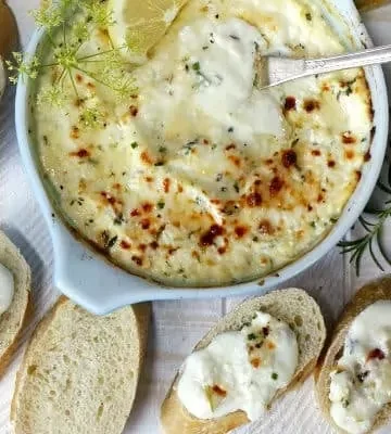 A bowl of creamy baked dip with herbs, accompanied by sliced bread, some topped with the dip. A sprig of rosemary is placed nearby.