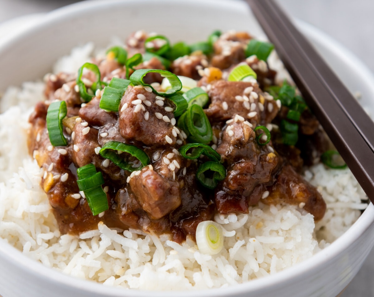A bowl of rice topped with diced meat in sauce, garnished with sesame seeds and sliced green onions, with chopsticks on the side.