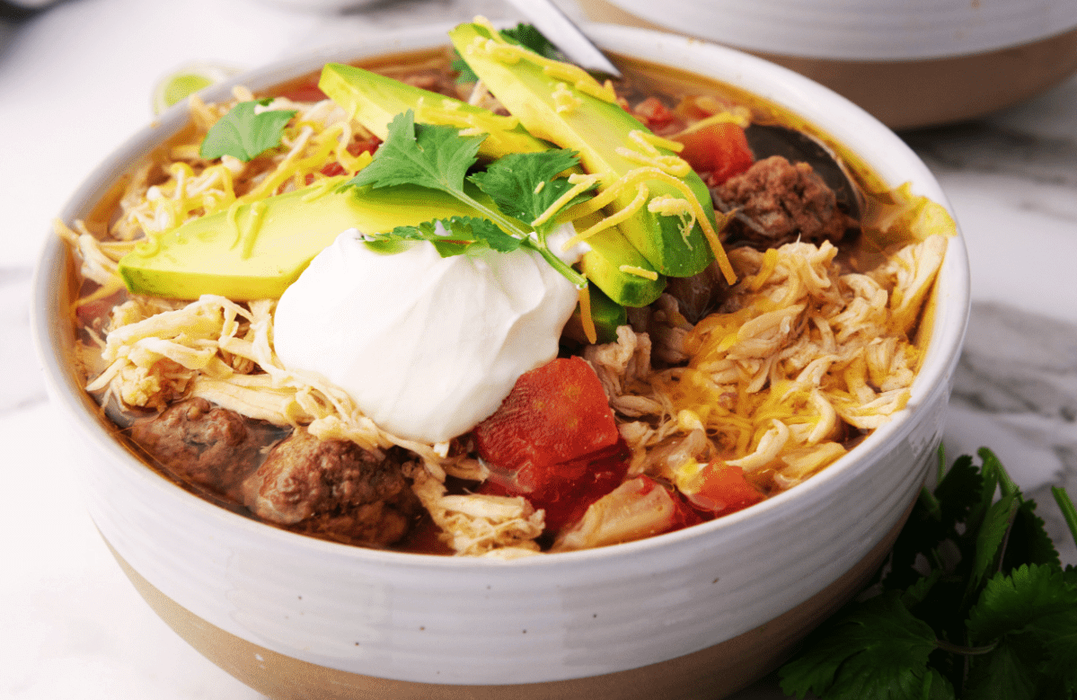 Bowl of soup topped with avocado slices, shredded cheese, sour cream, and cilantro. Contains shredded chicken, ground beef, and diced tomatoes.