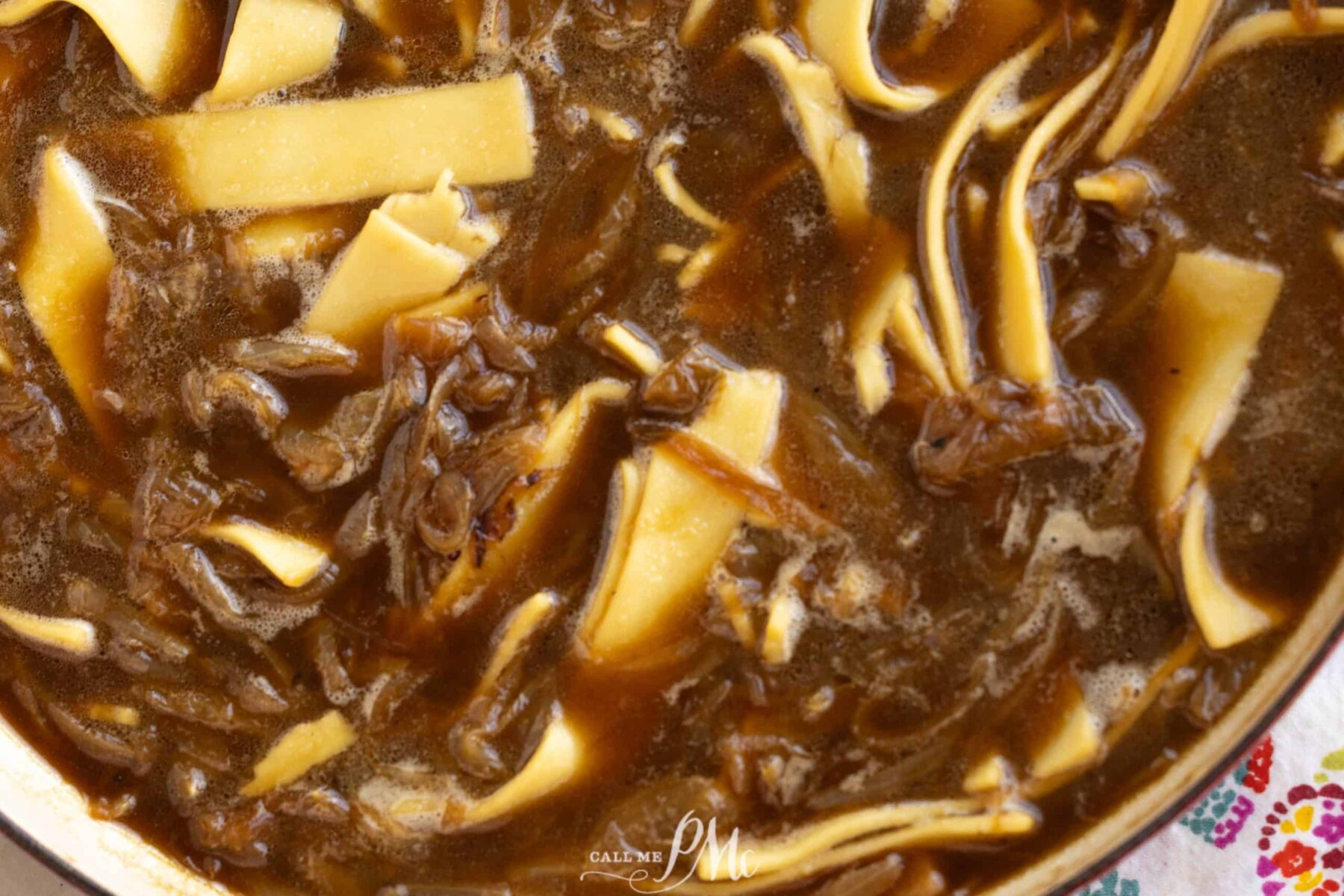 A close-up of a soup with dark broth, shredded meat, and wide noodle strips.