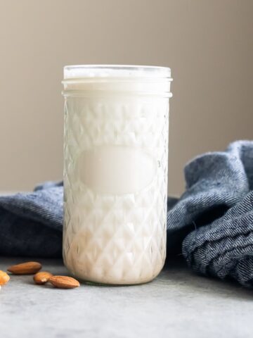 A clear glass jar filled with almond milk is placed on a gray surface, surrounded by a few almonds and a folded blue cloth.