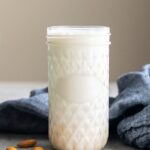 A glass jar filled with almond milk sits on a countertop beside a few almonds and a gray cloth.