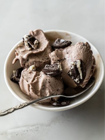 A bowl of chocolate ice cream with Oreo cookie pieces on top. A spoon rests in the bowl.