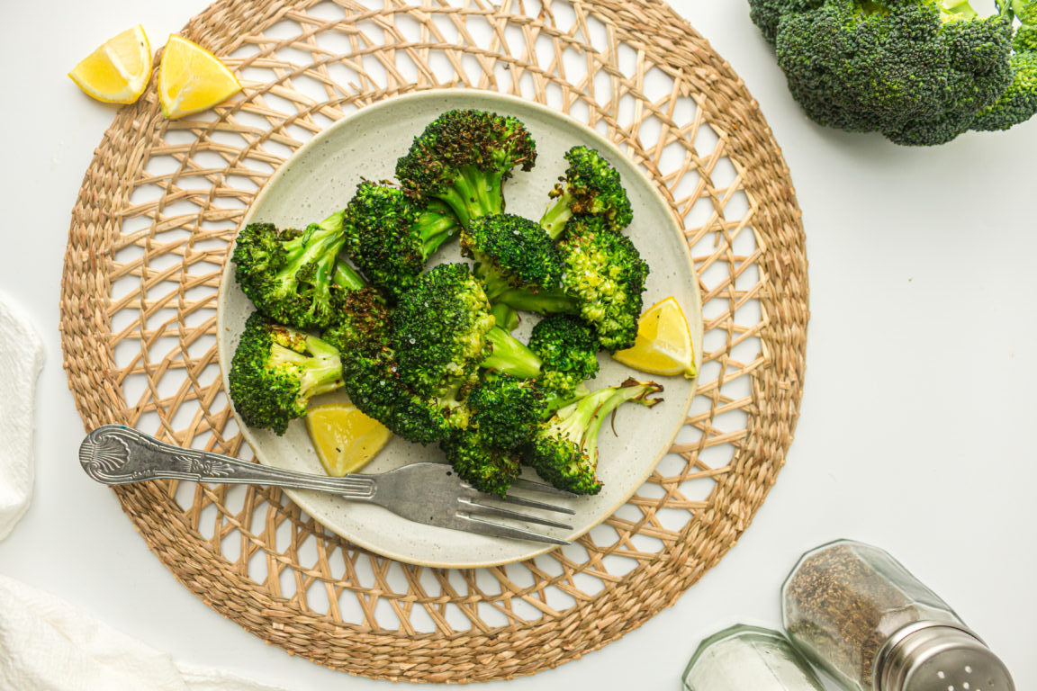 A plate of roasted broccoli with lemon wedges on a woven placemat. A fork rests on the plate. A whole broccoli and salt shaker are nearby.
