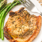 Grilled pork chop seasoned with herbs, served on a white plate with a side of cooked asparagus, fork placed next to the chop.