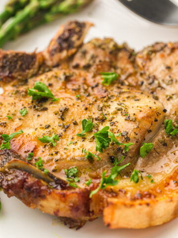 Cooked pork chop garnished with herbs, served on a white plate with asparagus in the background.