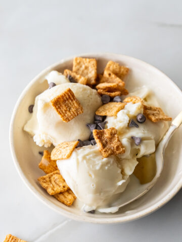 A bowl of vanilla ice cream topped with cereal squares and chocolate chips, with a spoon beside it on a light surface.