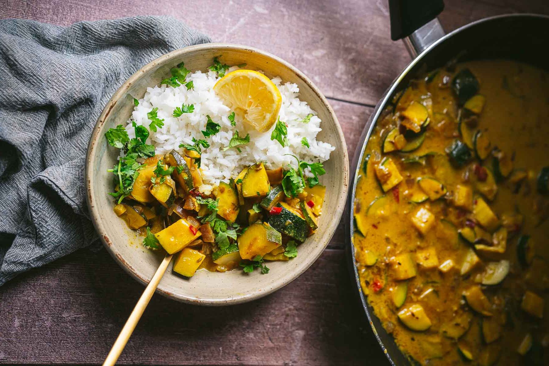 A bowl of rice with vegetable curry garnished with a lemon slice and cilantro, next to a pan containing more curry on a wooden table.