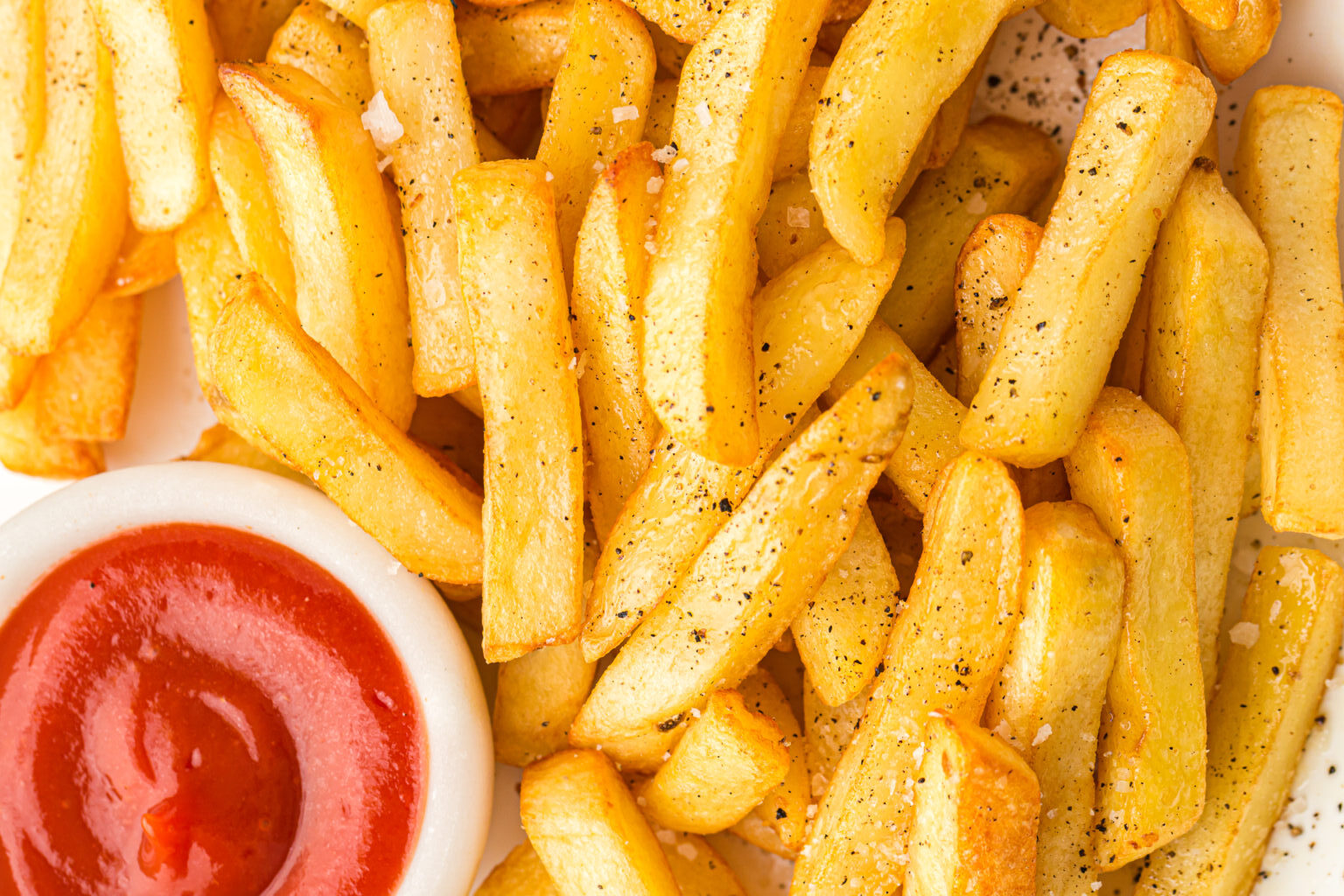 Close-up of golden French fries seasoned with salt and pepper, next to a small dish of ketchup.