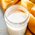 A glass jar filled with oat milk sits on a white surface next to scattered oats and an orange cloth.