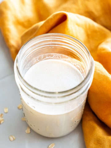 A glass jar filled with oat milk sits on a white surface next to scattered oats and an orange cloth.