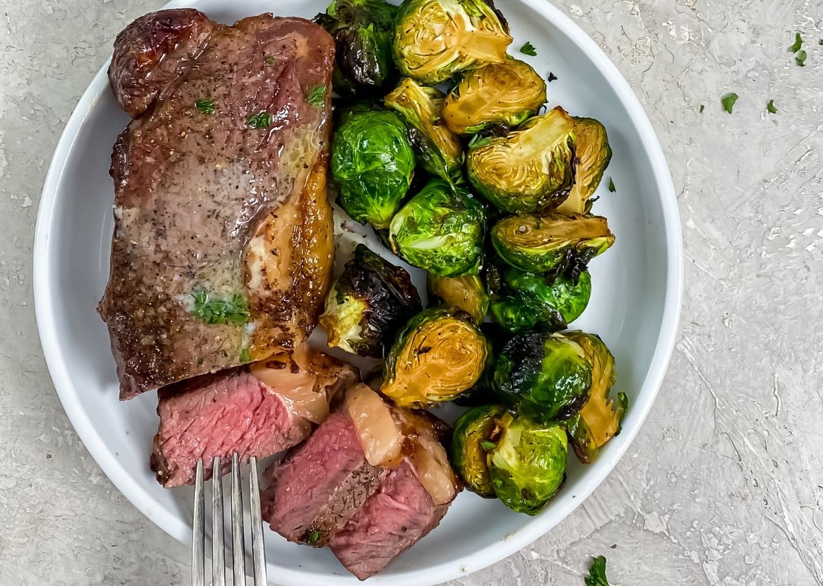 A plate with a cooked steak, some slices cut, and roasted Brussels sprouts.
