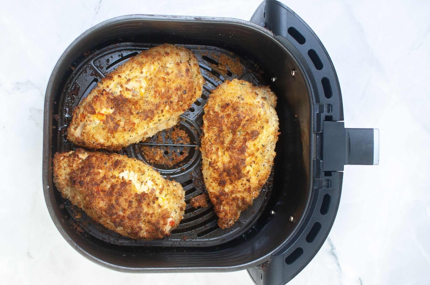 Three breaded chicken breasts in an air fryer basket on a white surface.