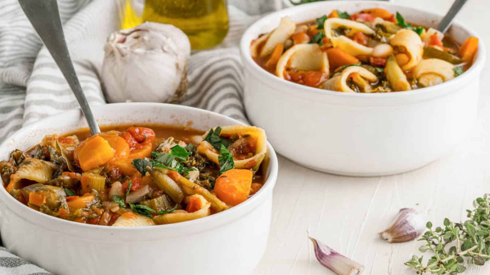 Two bowls of vegetable pasta soup with spoons on a white surface. Garlic, a striped cloth, and a bottle of olive oil in the background.