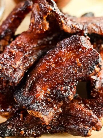 A pile of cooked, glazed pork ribs on a wooden surface, with a small bowl of sauce in the background.