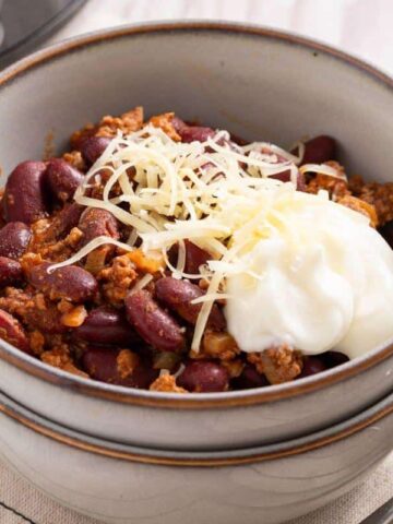 A bowl of chili with kidney beans and ground meat, topped with shredded cheese and sour cream, on a napkin beside two spoons.