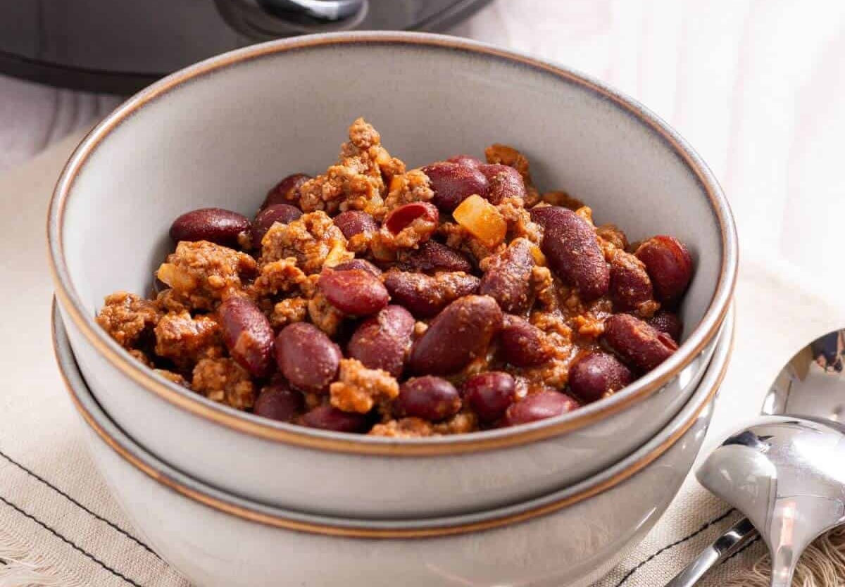 A bowl of chili with kidney beans and ground meat, placed on a stack of bowls next to a spoon on a table.
