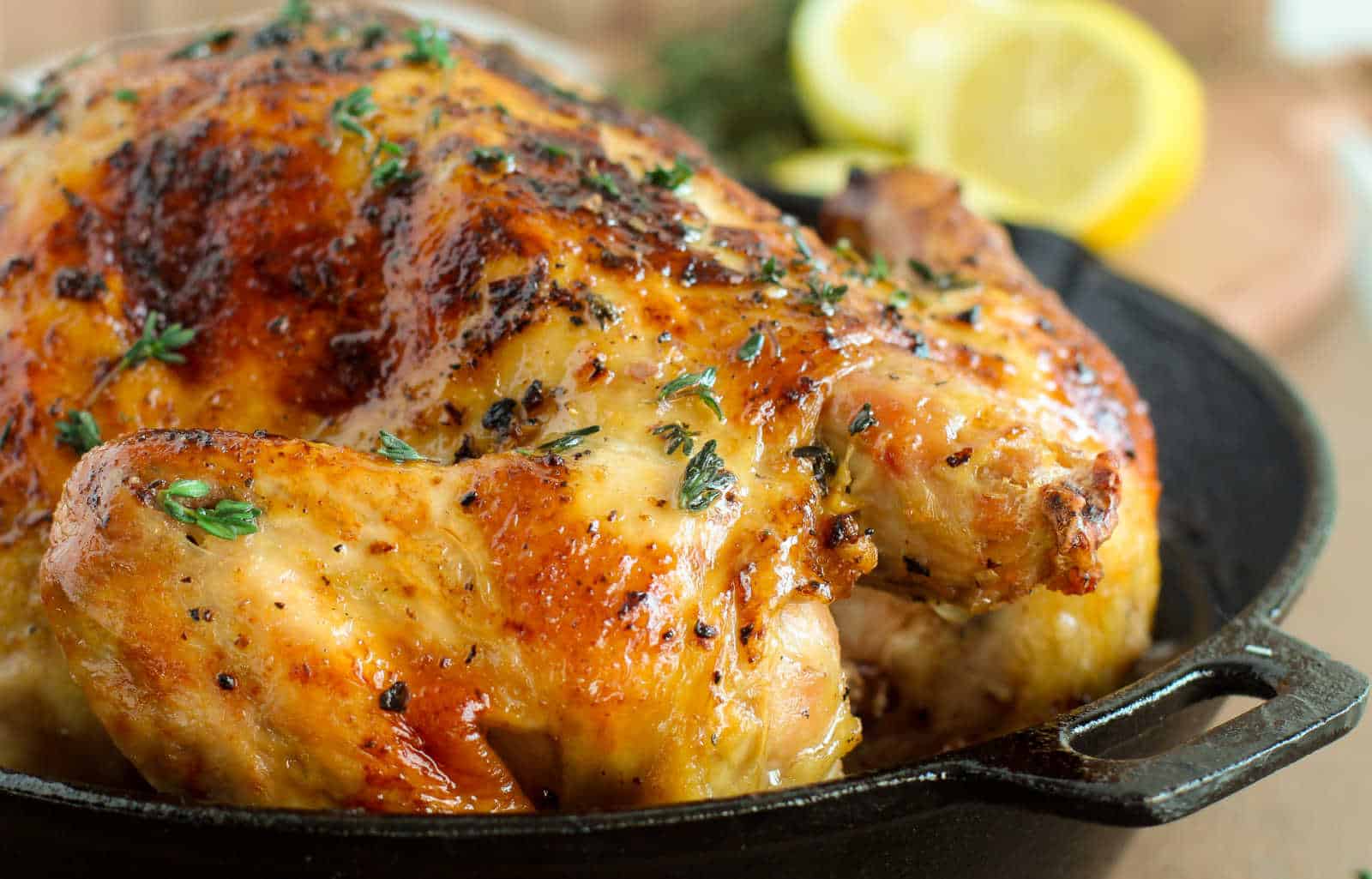 Roasted whole chicken in a skillet, garnished with herbs, with lemon slices in the background.