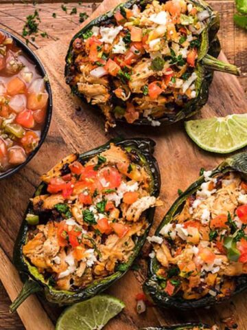 Three stuffed poblano peppers filled with chicken, cheese, and vegetables on a wooden board. A bowl of salsa and lime wedges are on the side.