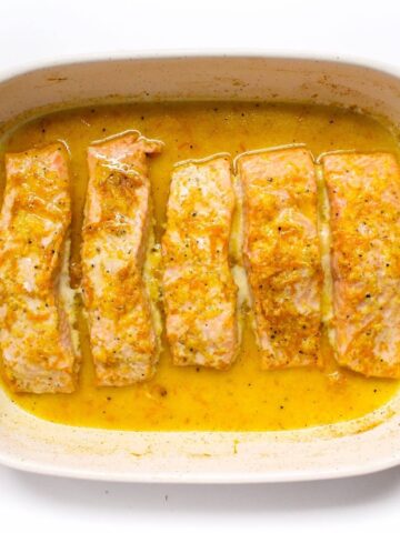 Five salmon fillets in a creamy, orange-hued sauce in a white baking dish on a wooden board.