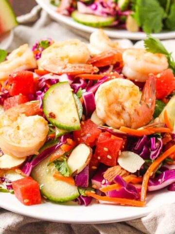 A plate of shrimp salad with sliced cucumbers, red cabbage, carrots, watermelon, almonds, and cilantro, served on a white plate.