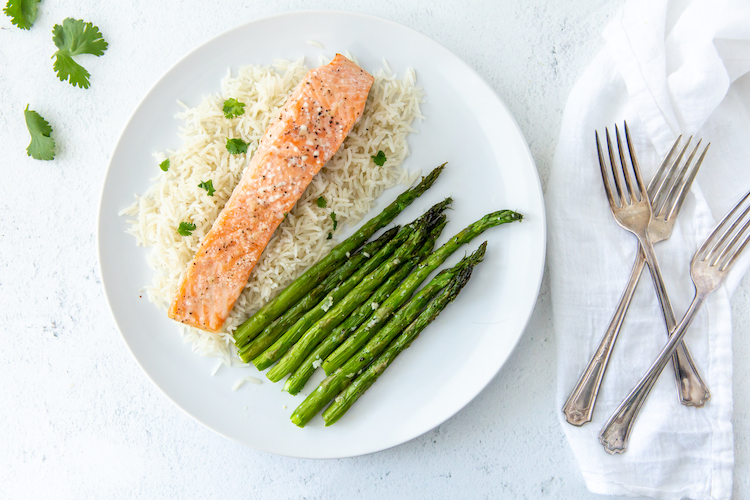 A plate with cooked salmon, white rice, and asparagus makes for a perfect easy dinner. Silver forks rest on a white cloth next to the plate, while a few cilantro leaves are scattered nearby.