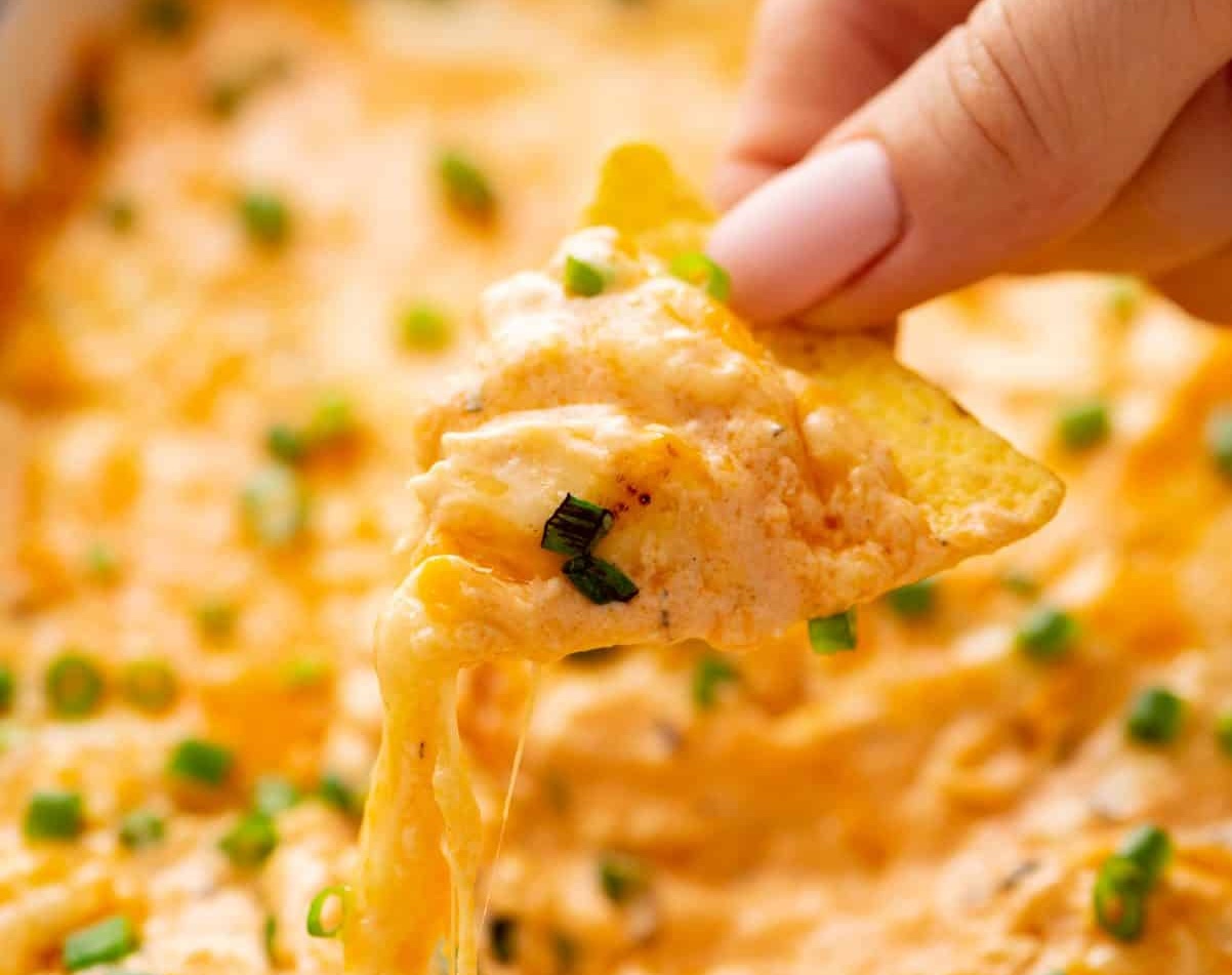 Close-up of a hand holding a tortilla chip dipped in cheesy dip with green onions, showing a string of melted cheese.