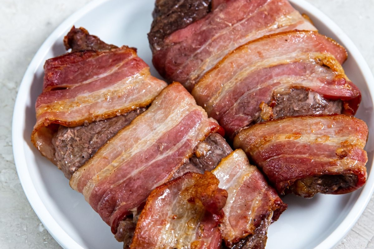 Two pieces of cooked meat wrapped in crispy bacon, placed on a white plate.