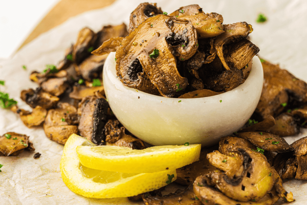 Sautéed mushroom slices in a small white bowl, garnished with chopped herbs, with lemon wedges on the side.