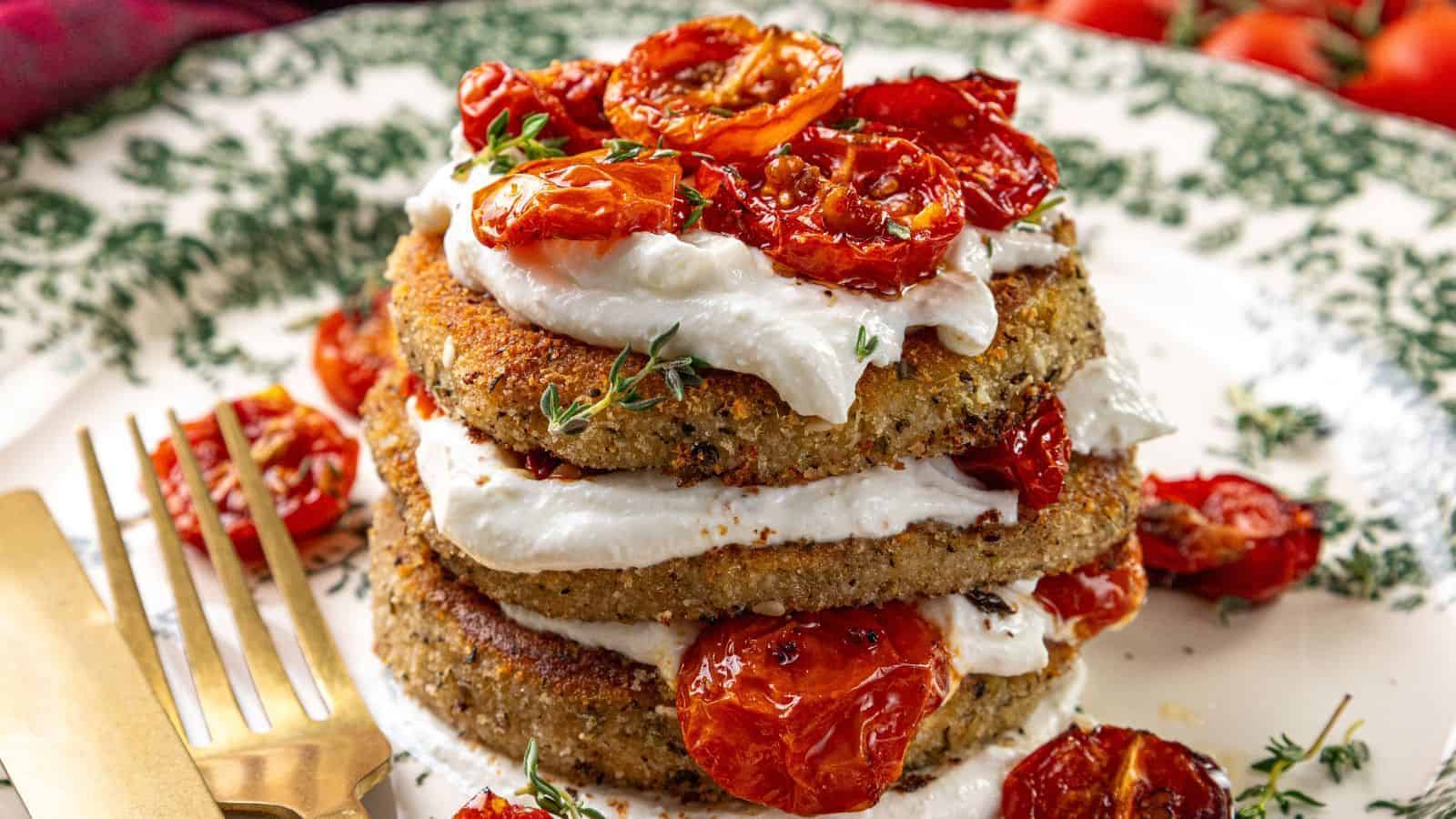 Stack of breaded eggplant slices layered with whipped feta and roasted tomatoes on a decorative plate, with a fork and knife on the side.