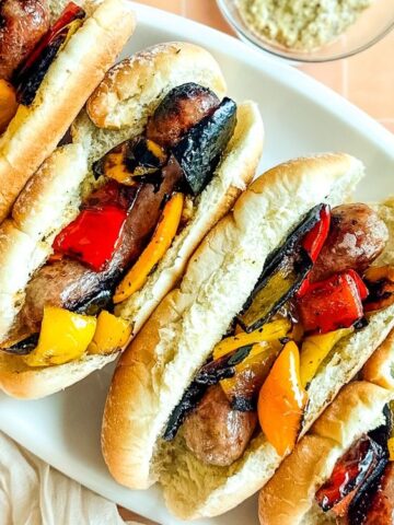 Four sausage sandwiches in buns topped with grilled red, yellow, and orange bell peppers on a white platter, with small bowls of ketchup and mustard nearby.