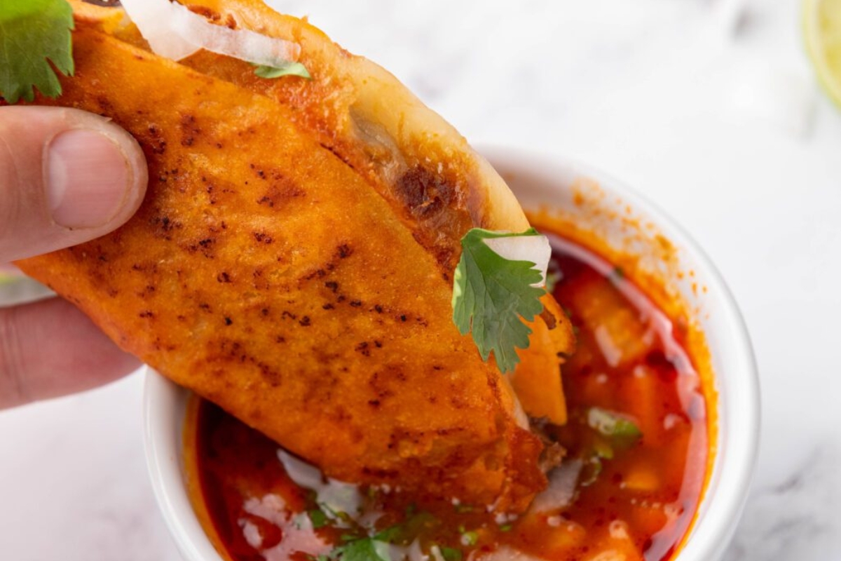 A hand holds a crispy taco filled with meat and onions, dipping it into a cup of red consommé garnished with cilantro.