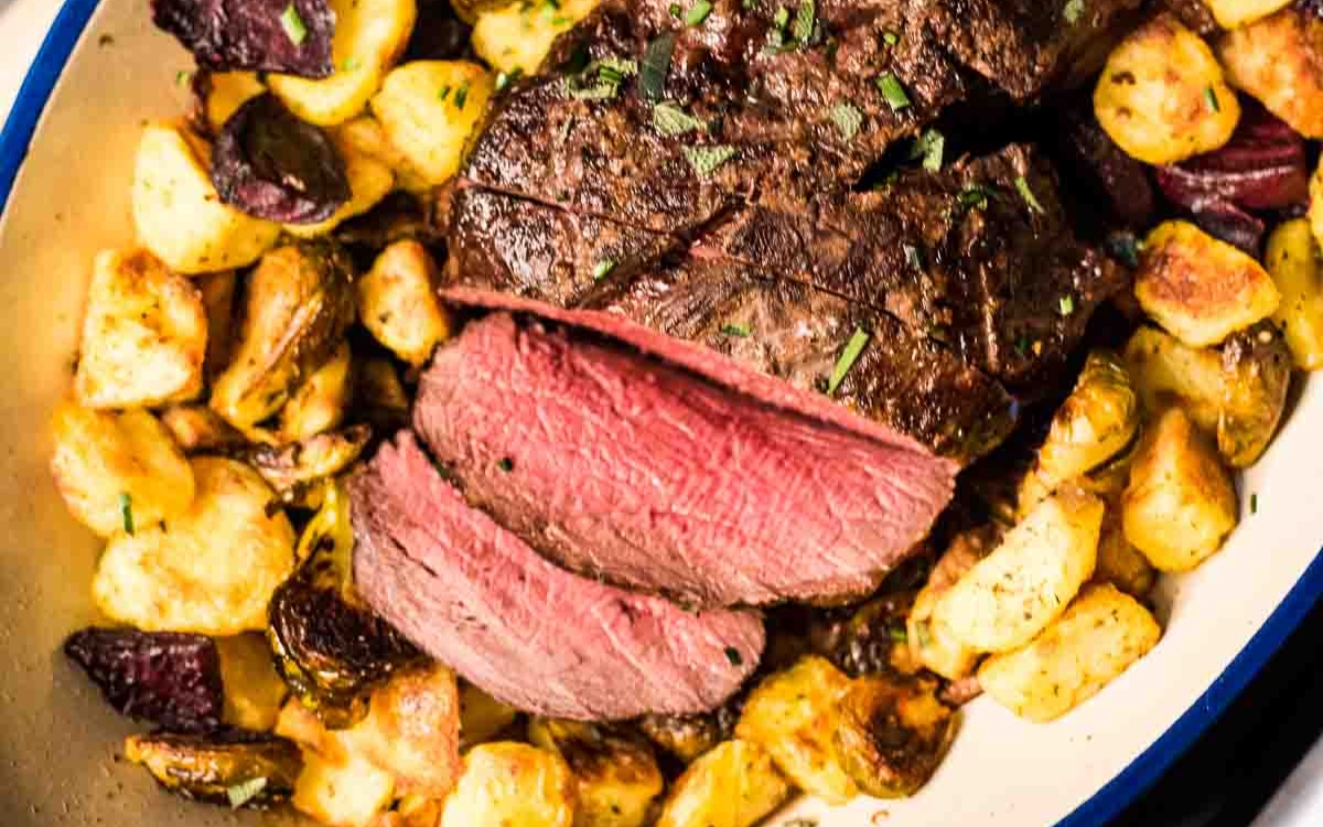 Sliced medium-rare beef served on a bed of roasted potatoes and vegetables, garnished with chopped herbs.