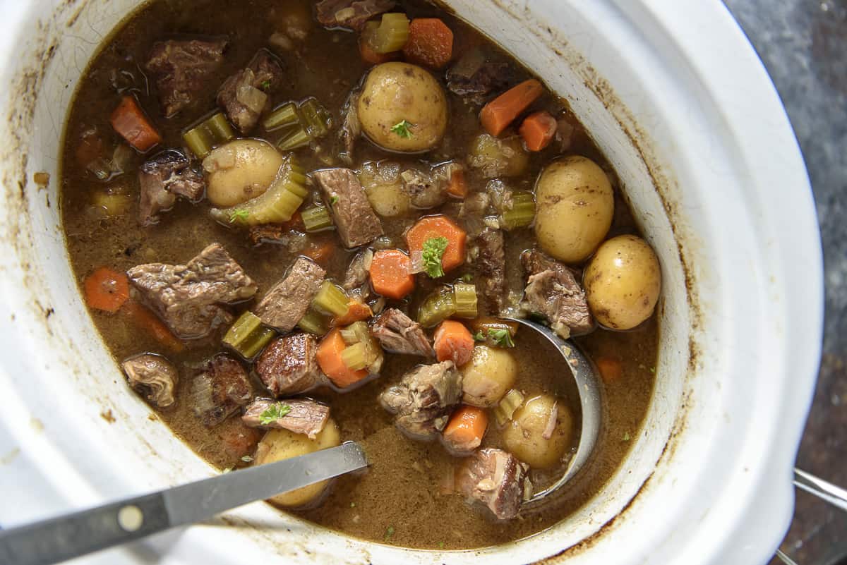 A slow cooker filled with beef stew, featuring chunks of beef, carrots, celery, and baby potatoes in a brown broth. A ladle and a serving utensil are visible in the stew.