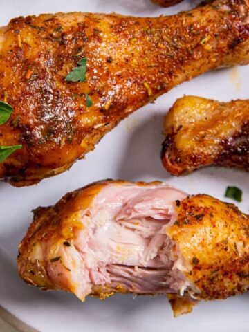 Three cooked chicken drumsticks with seasoning on a white plate, with one drumstick partially eaten, garnished with small parsley leaves.