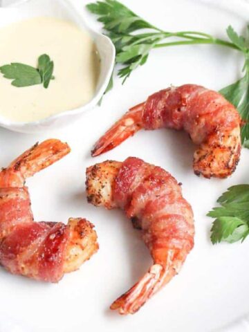 Three bacon-wrapped shrimp are elegantly arranged on a white plate, accented with parsley and served alongside a small bowl of dipping sauce. These mouthwatering appetizers promise to delight your taste buds.