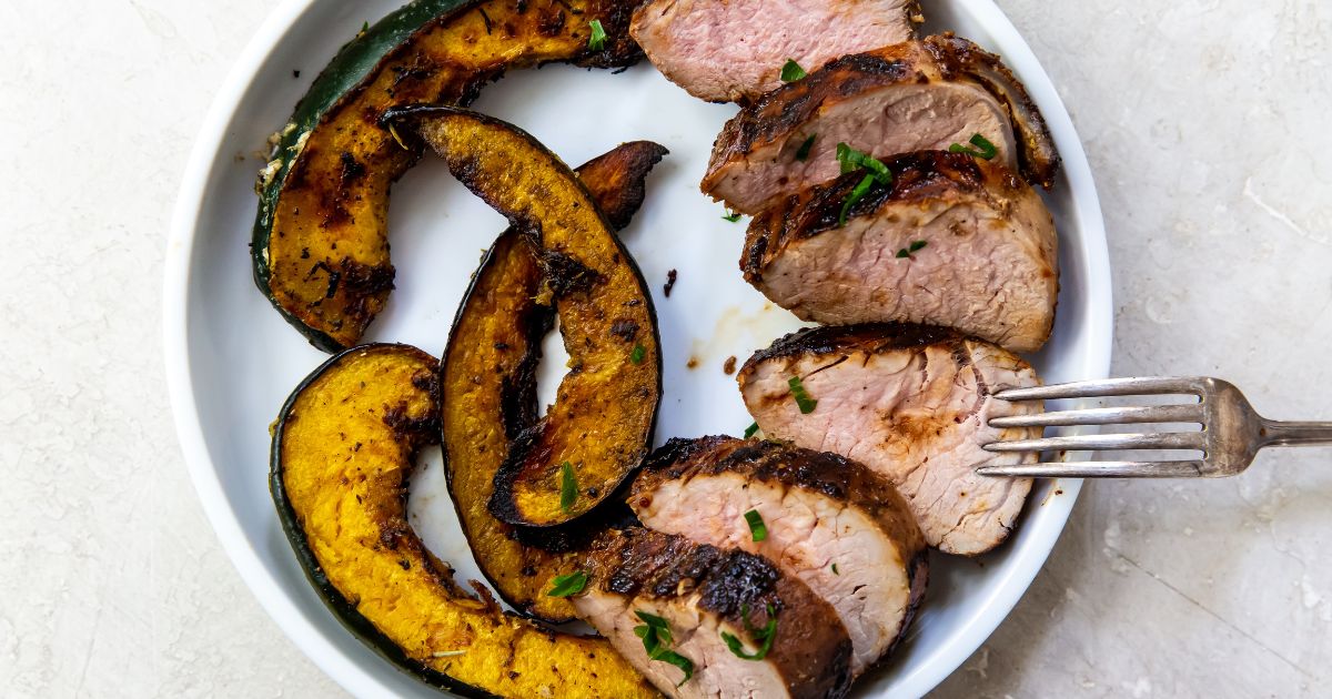 A white plate with sliced roasted pork tenderloin and roasted acorn squash, with a fork resting on the pork.
