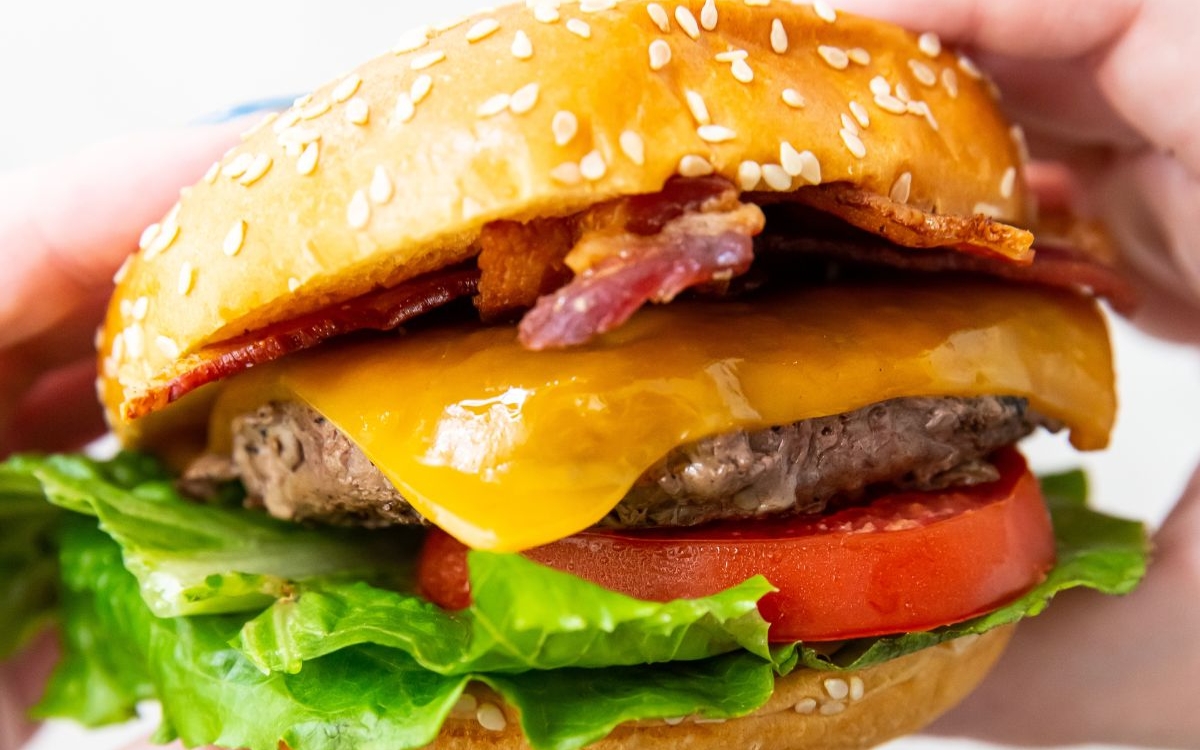 A close-up of a cheeseburger with lettuce, tomato, cheddar cheese, bacon, and a beef patty in a sesame seed bun.