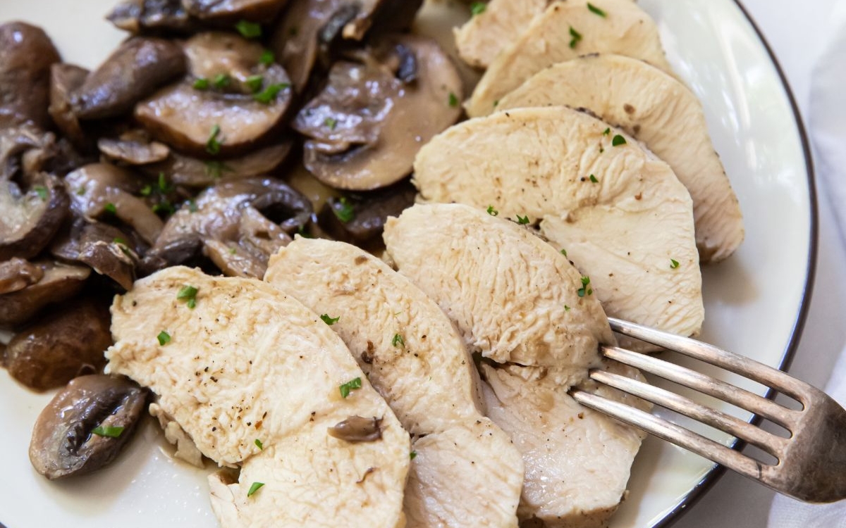 Sliced cooked chicken breast and sautéed mushrooms on a white plate with a fork.