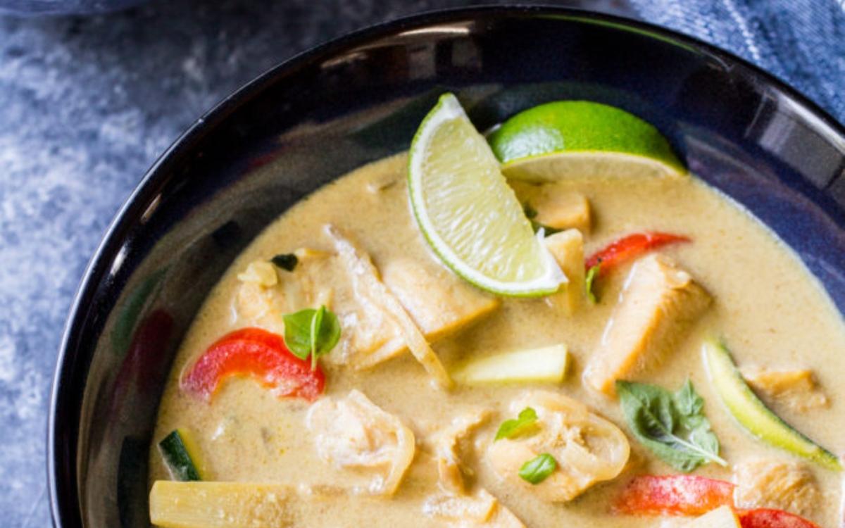 A bowl of Thai green curry with chicken, red bell pepper, zucchini, bamboo shoots, fresh basil, and a lime wedge.
