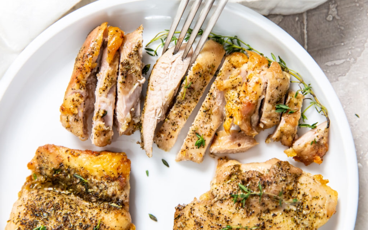 A white plate with sliced, seasoned cooked chicken thighs, garnished with fresh thyme. A fork is holding one chicken slice.