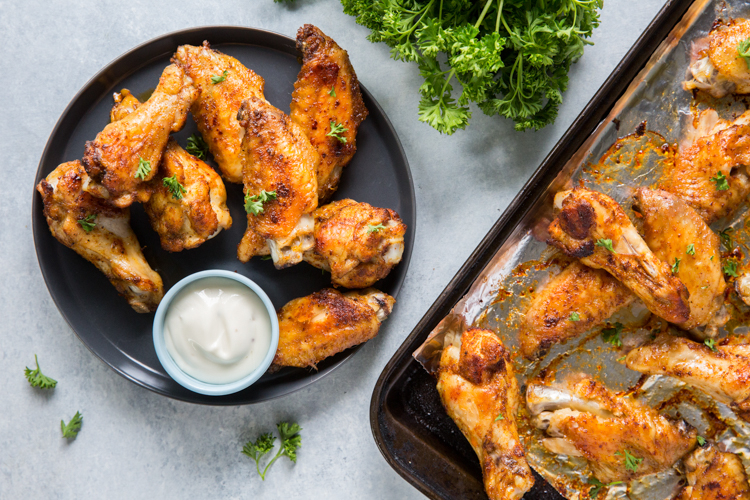A black plate with baked chicken wings and a cup of ranch dip, next to a baking tray with more wings and sprigs of parsley.