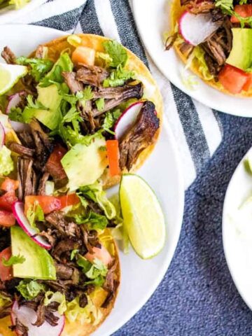 Three plates of shredded beef tostadas topped with avocado, tomato, lettuce, cilantro, radish, and lime wedges are arranged on a striped cloth—perfect inspiration for your next taco recipes.