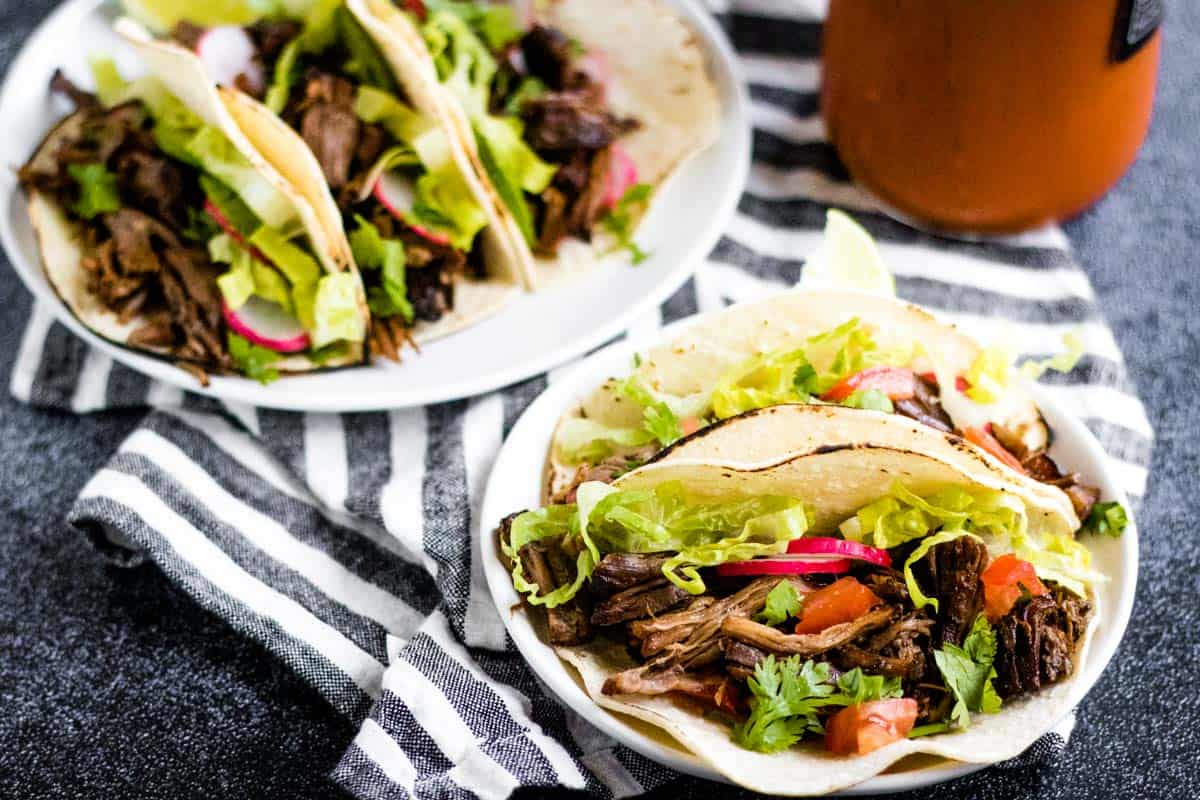 Two plates of beef tacos with shredded meat, lettuce, cilantro, radish, and tomato on corn tortillas showcase classic taco recipes, beautifully arranged on a striped cloth.