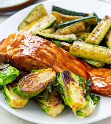 A plate with glazed salmon, roasted Brussels sprouts, and seasoned zucchini slices on a white surface.